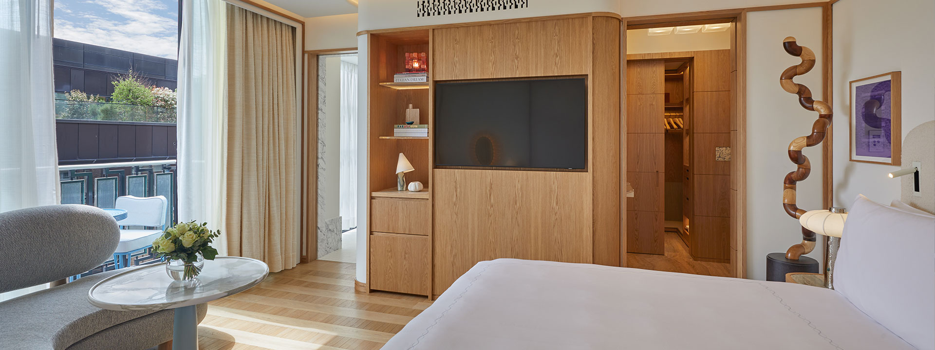 Edge of bed with white bedding in bedroom with wooden walk in wardrobe, TV on wall and open glass doors leading to balcony